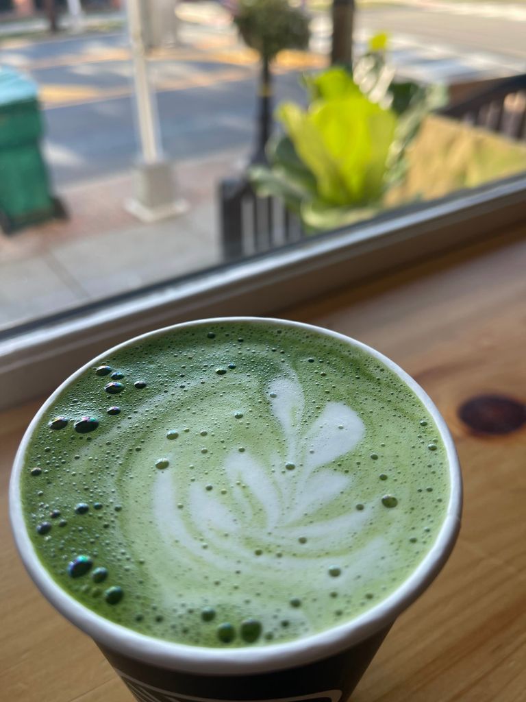 A cup of green matcha tea with a decorative foam design, set on a wooden table with a view of a street and greenery in the background.