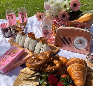 A picnic spread featuring pink drinks, colorful snacks, fresh fruits, and a vintage radio, surrounded by flowers on a blanket in a sunny park.