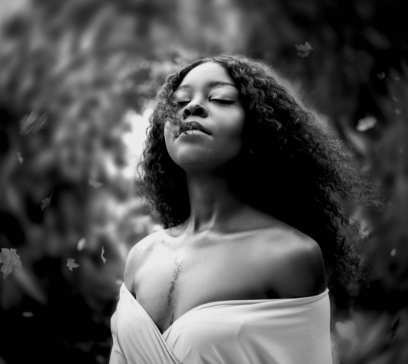 A serene Black woman with curly hair, wearing an off-shoulder white dress, stands outdoors with her eyes closed, enjoying a moment of peace amidst soft, blurred greenery.
