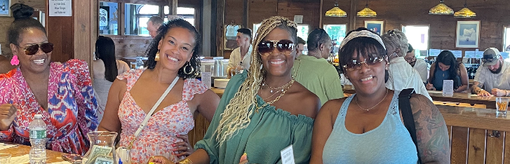 A group of four women smiling and posing together at a restaurant, wearing stylish summer outfits and sunglasses, with a lively atmosphere in the background.