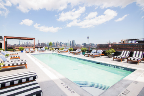 A rooftop pool area featuring striped lounge chairs, surrounded by potted plants, with a skyline view of the city and blue sky.