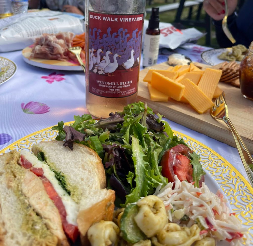 A delicious meal featuring a sandwich with tomato and greens, a fresh salad, pasta salad, cheddar cheese, and a bottle of Duck Walk Vineyards Windmill Blush wine, set on a floral tablecloth.