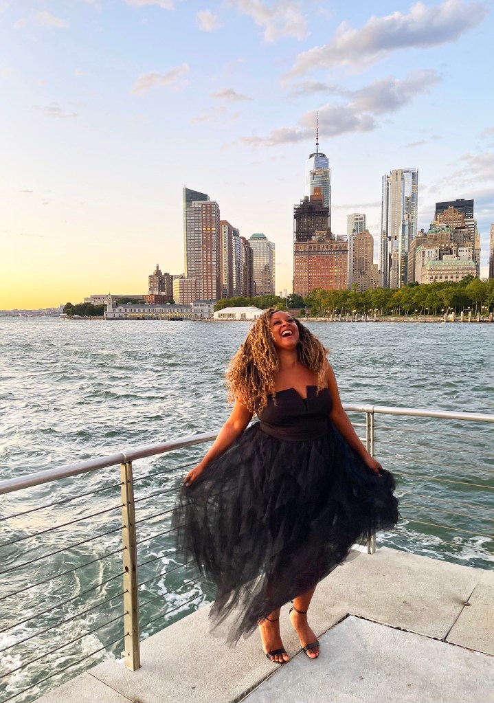 A joyful woman in a black tulle dress stands near the water, with a city skyline in the background during sunset.