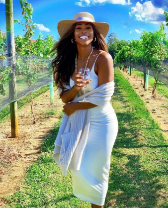 A smiling woman in a white dress and wide-brimmed hat stands in a vineyard, holding a glass of wine with lush vines and a blue sky in the background.