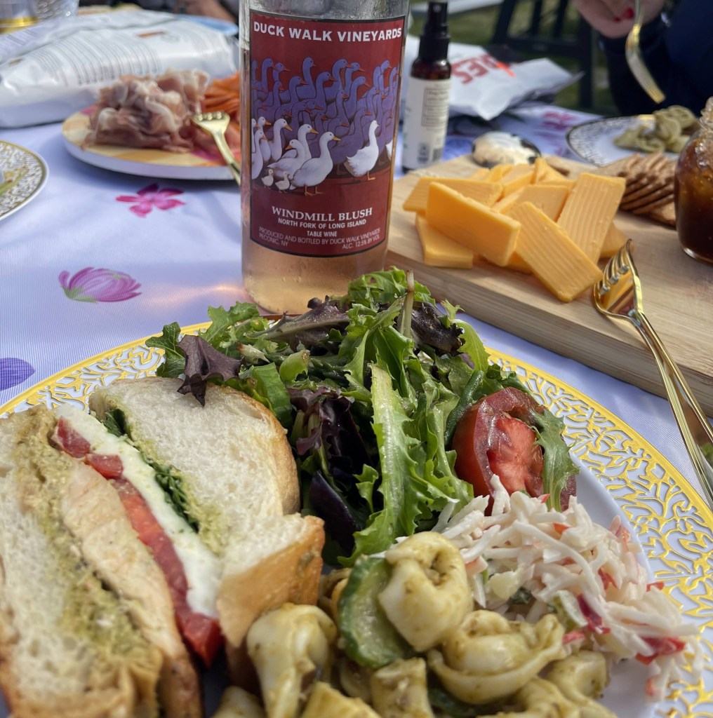 A plate featuring a sandwich with chicken, pesto, and tomato, accompanied by a mixed green salad, coleslaw, and tortellini pasta, alongside a bottle of Windmill Blush wine and a cheese platter with crackers.