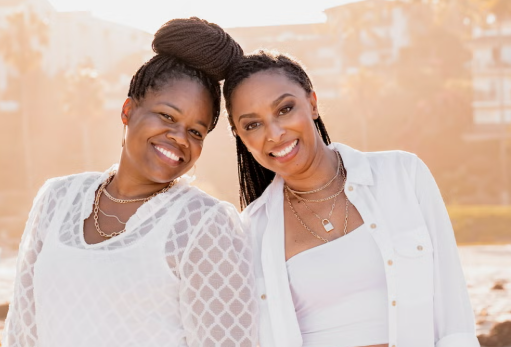 Two smiling Black women standing close together, dressed in casual, stylish attire, with a coastal background illuminated by warm sunlight.