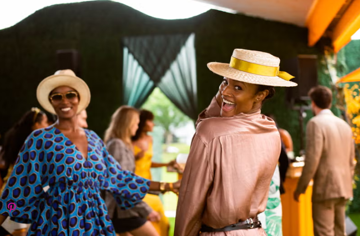 Two Black women joyfully dancing at a vibrant outdoor gathering, wearing fashionable outfits and hats.