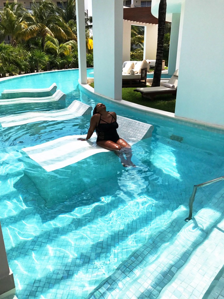 A person relaxing in a pool, lying on a partially submerged platform, surrounded by palm trees and a serene atmosphere.