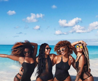 Four Black women posing together on a beach, wearing stylish black swimsuits and sunglasses, with the ocean and blue sky in the background.