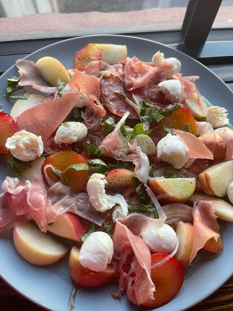 A vibrant summer salad featuring sliced peaches, apples, fresh mozzarella, and prosciutto, garnished with herbs, arranged on a gray plate near a window.