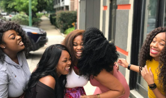 Five Black women joyfully laughing and embracing each other outdoors, showcasing connection and friendship.