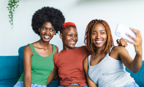 Three Black women smiling and taking a selfie together, showcasing friendship and connection in a cozy indoor setting.