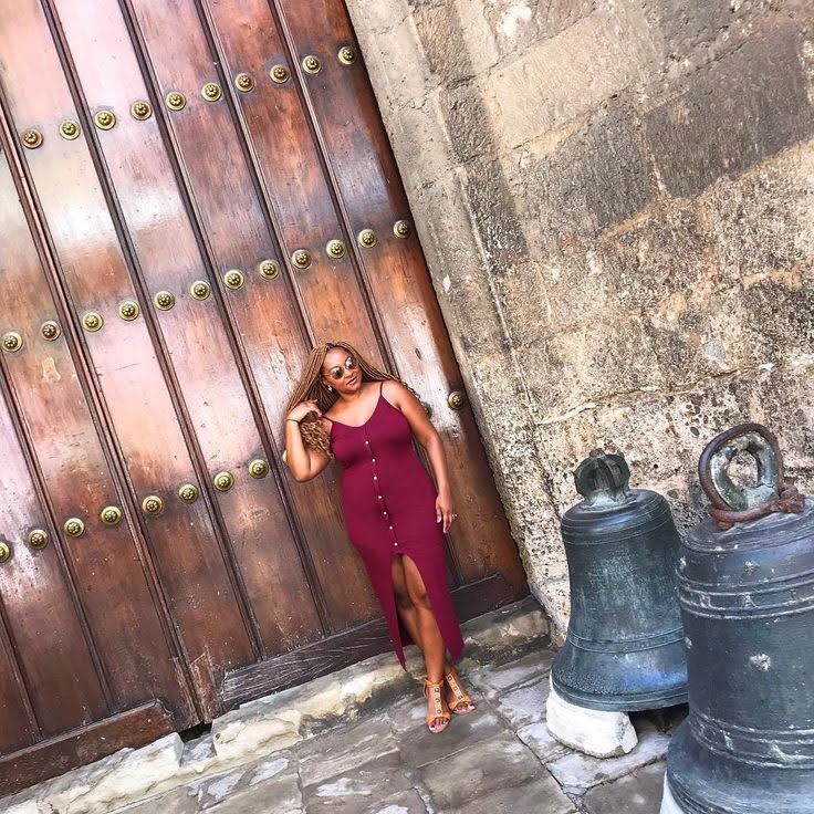 A woman in a burgundy dress poses next to a large wooden door with metal knobs, with bells in the foreground.