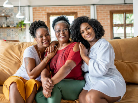 Three Black women sitting together on a couch, smiling and embracing each other in a warm, inviting setting.