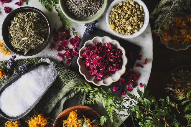 A collection of dried herbs and flowers arranged on a marble surface, including lavender, chamomile, and rose petals, alongside a small bowl of salt.