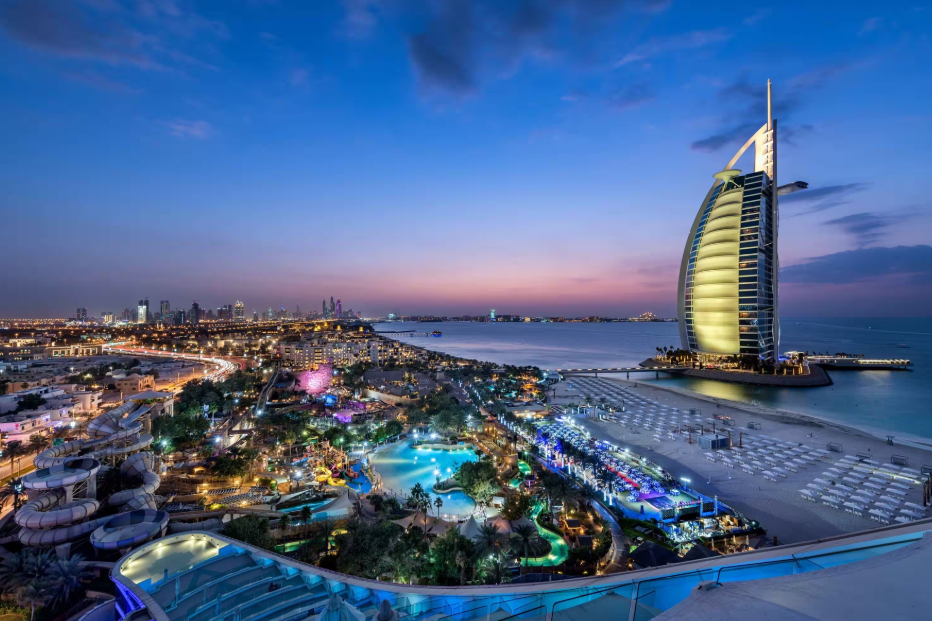 Panoramic view of Dubai's coastline featuring the iconic Burj Al Arab at sunset, showcasing a beautifully illuminated cityscape and beach area.