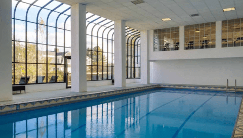 Indoor swimming pool with large windows allowing natural light, featuring a clear blue water surface and seating area beside the pool.