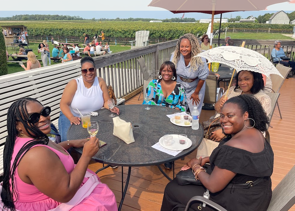 A group of Black women enjoying a wine tasting on a wooden deck, with a vineyard in the background. They are smiling and holding glasses of wine, showcasing a joyful and festive atmosphere.
