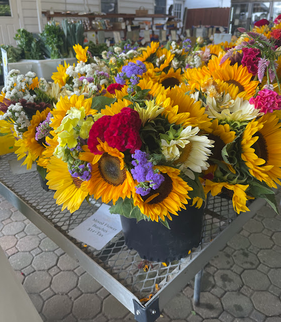 A vibrant display of flower bouquets featuring sunflowers, colorful blooms, and greenery, arranged on a table with a price sign in a market setting.