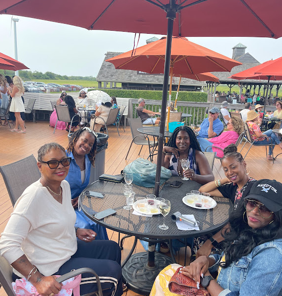 A group of Black women enjoying a wine tasting on a patio in the Hamptons, seated at a round table with glasses of wine and food. Bright umbrellas provide shade in the background.