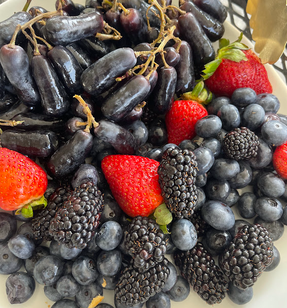 A vibrant assortment of fresh berries including strawberries, blueberries, blackberries, and a unique variety of small dark grapes arranged on a plate.
