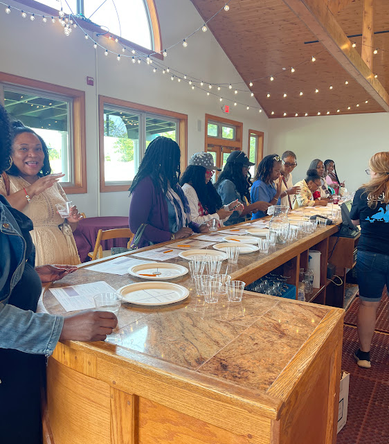 A group of diverse women enjoying a wine tasting event at a wooden bar, with glasses and plates in front of them. The atmosphere is lively, featuring string lights and large windows.