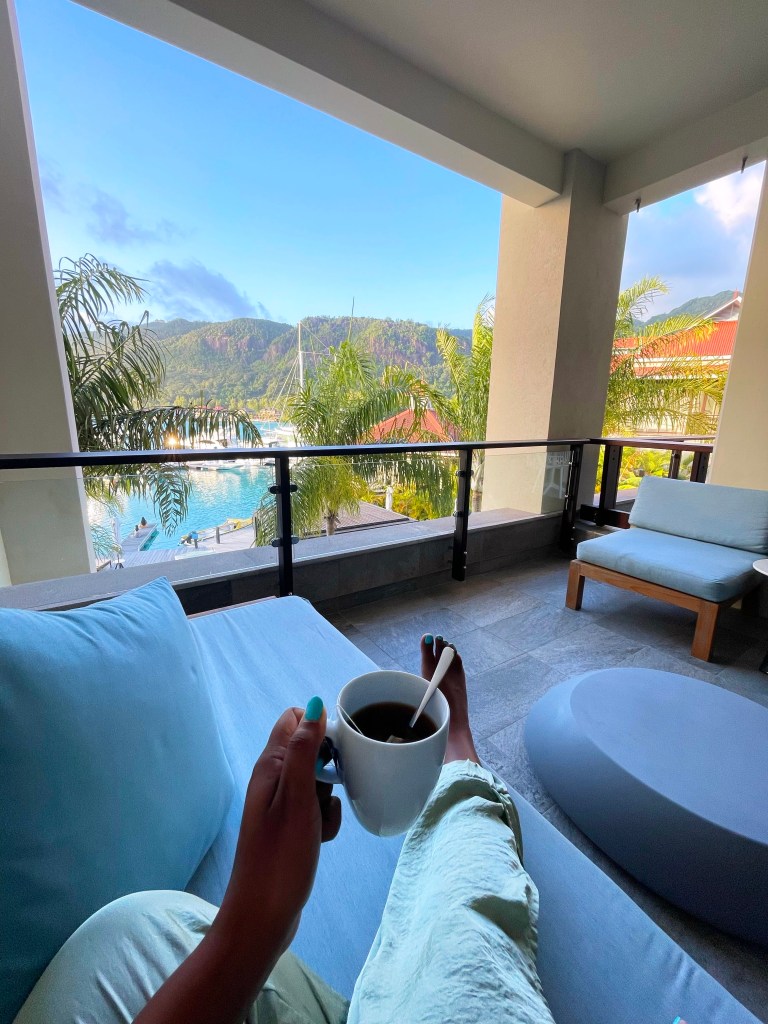 A person relaxes on a balcony with light-colored furnishings, holding a cup of coffee while overlooking a scenic view of a harbor and lush mountains in the distance.