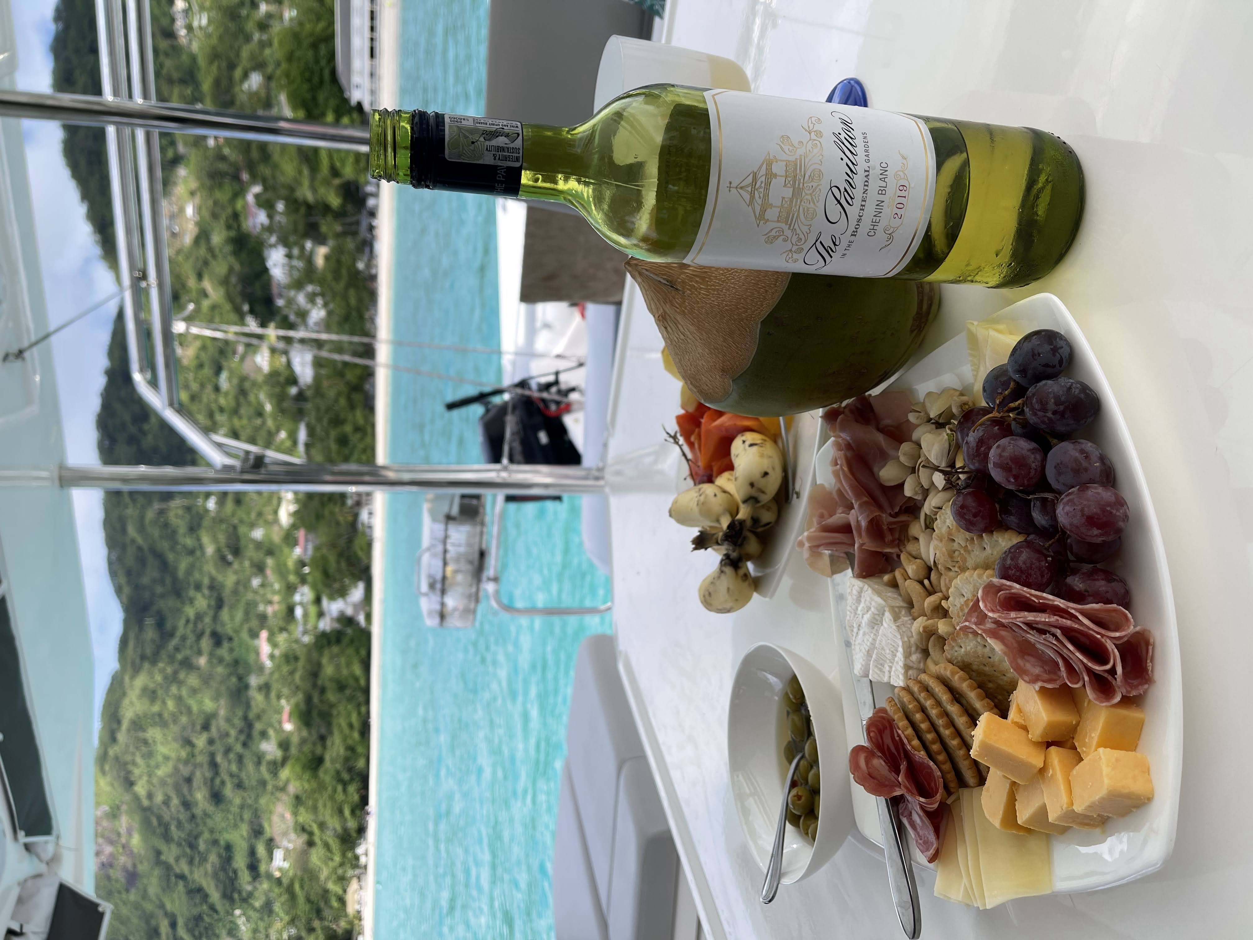 A beautifully arranged plate of charcuterie with cheese, crackers, grapes, and cured meats, accompanied by a bottle of white wine and a coconut, set against a backdrop of turquoise water and lush green hills.